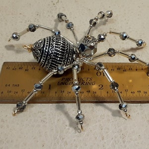 May include: A silver and black beaded spider with a decorative, patterned body. The spider is approximately 2 inches long and is sitting on a ruler.