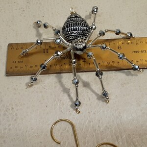 May include: A silver and black beaded spider ornament with a decorative metal body. The spider has a gold hanger attached to the top.