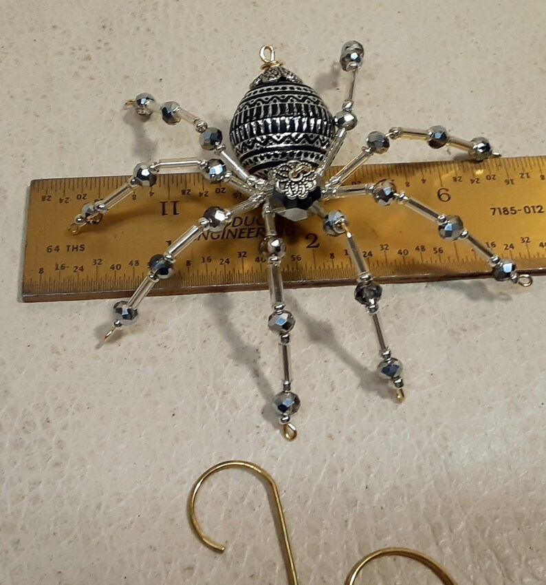 May include: A silver and black beaded spider ornament with a decorative metal body. The spider has a gold hanger attached to the top.