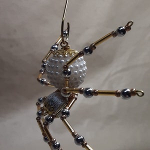 May include: A handcrafted spider ornament with a white beaded body and a silver and gold metallic body. The spider's legs are made of gold-colored rods and silver beads. A gold wire loop is attached for hanging.