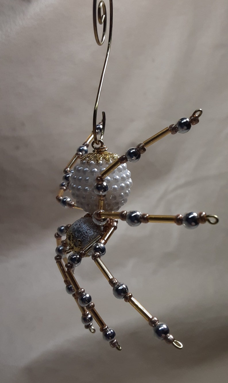 May include: A handcrafted spider ornament with a white beaded body and a silver and gold metallic body. The spider's legs are made of gold-colored rods and silver beads. A gold wire loop is attached for hanging.