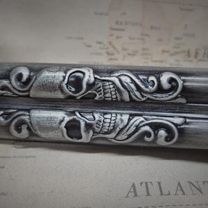 May include: Two silver-toned metal skull and floral designs on a black background. The skulls have a detailed design with a floral pattern on each side.