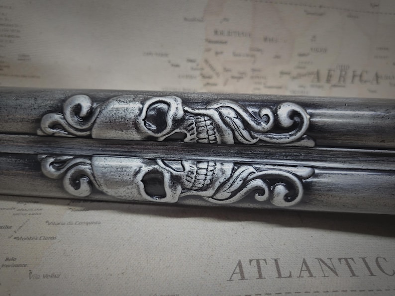 May include: Two silver-toned metal skull and floral designs on a black background. The skulls have a detailed design with a floral pattern on each side.