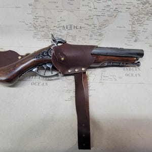 May include: A brown leather holster with a metal buckle and a brown wooden pistol with a silver barrel and decorative engravings. The holster is attached to a brown leather strap.