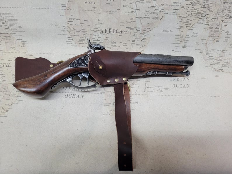 May include: A brown leather holster with a metal buckle and a brown wooden pistol with a silver barrel and decorative engravings. The holster is attached to a brown leather strap.