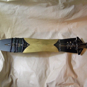 May include: A black and gold spear with a leather wrap and a pointed tip. The spear is made of metal and has a decorative design.