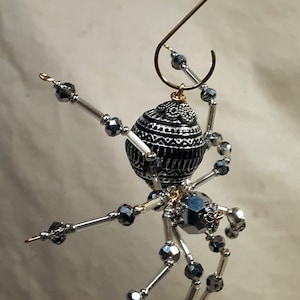 May include: A silver and black beaded spider ornament with a gold hanger. The spider's body is a round, black bead with silver accents. The legs are made of silver wire and beads.