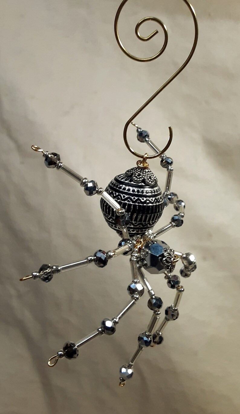 May include: A silver and black beaded spider ornament with a gold hanger. The spider's body is a round, black bead with silver accents. The legs are made of silver wire and beads.