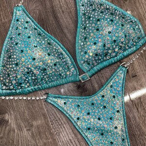 May include: Teal and silver sequined bikini with crystal embellishments. The bikini top has a halter style with a clasp closure in the back. The bikini bottom has a high-cut style.