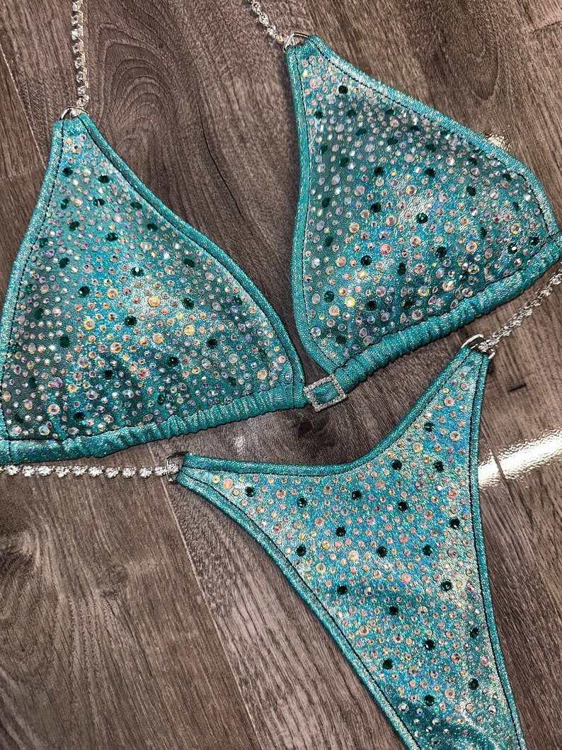 May include: Teal and silver sequined bikini with crystal embellishments. The bikini top has a halter style with a clasp closure in the back. The bikini bottom has a high-cut style.