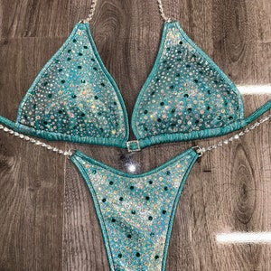May include: A teal blue bikini with crystal embellishments. The bikini top has a halter style with a clasp closure in the back. The bikini bottom has a high-cut style.