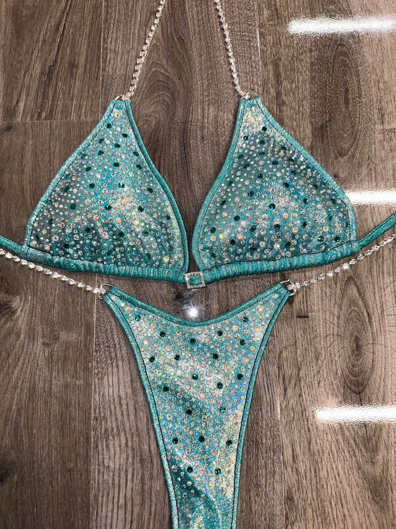 May include: A teal blue bikini with crystal embellishments. The bikini top has a halter style with a clasp closure in the back. The bikini bottom has a high-cut style.