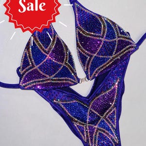 May include: A purple and blue sequined competition bikini with a geometric design. The bikini is decorated with rhinestones and has a v-neckline.