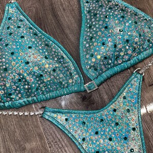 May include: Teal and silver sequined bikini with crystal embellishments. The bikini top is a triangle style with a clasp closure. The bikini bottom is a high-cut style.