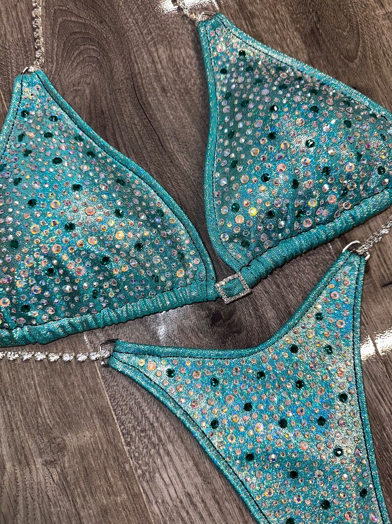 May include: Teal and silver sequined bikini with crystal embellishments. The bikini top is a triangle style with a clasp closure. The bikini bottom is a high-cut style.