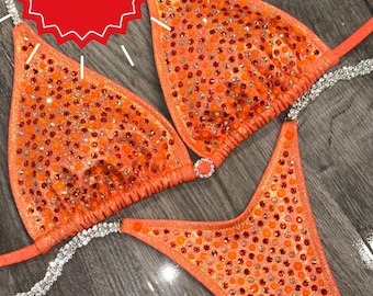 Puede incluir: Un bikini de color coral con un diseño incrustado de cristales. La parte superior del bikini tiene forma triangular y la parte inferior es de estilo de corte alto. El bikini está sobre una superficie de madera.