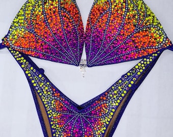 May include: A purple bikini with a colorful crystal design. The top and bottom of the bikini feature a gradient of orange, pink, yellow, and green crystals.