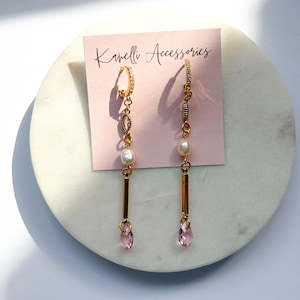 Peut inclure: Boucles d'oreilles pendantes dorées avec une gemme rose en forme de goutte d'eau en bas. Les boucles d'oreilles comportent une perle et une barre verticale dorée. Les boucles d'oreilles sont présentées sur une carte rose avec "Kawelli Accessories".