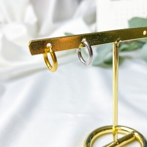 May include: A gold earring display stand with two hoop earrings. One earring is gold and the other is silver.