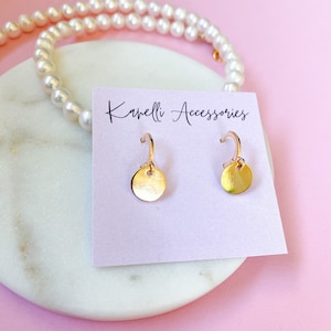 Op de afbeelding: Gouden creolen met een klein rond schijfje. De oorbellen worden gepresenteerd op een witte kaart met de tekst "Kanelli Accessories".