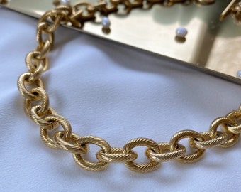 May include: A gold-toned chain necklace featuring large, oval-shaped links with a textured, rope-like finish. The necklace is constructed from interconnected metal links that form a continuous, heavy-looking chain design with a polished appearance.