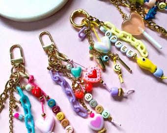 May include: Four gold-tone metal keychains featuring colorful plastic chain links, heart-shaped beads, and letter beads spelling out QUACK, CUTE, and COFFEE. The keychains include various charms like a pink chocolate bar, a lollipop, and smiley faces.