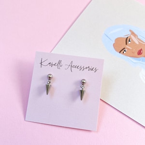 Può includere: Un paio di orecchini a punta argentati con una piccola sfera in cima a ogni punta. Gli orecchini sono esposti su una carta rosa con il testo "Kamelli Accessories".