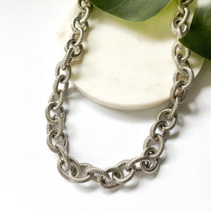 Può includere: Collana a catena color argento con un design testurizzato a forma di corda. La collana è esposta su una superficie bianca con una foglia verde sullo sfondo. Le maglie della catena sono spesse e ravvicinate.