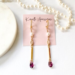 May include: A pair of gold-tone drop earrings featuring a small hoop with clear stones, a white pearl, a marquise-cut clear stone, a gold bar, and a deep pink teardrop crystal. The earrings are displayed on a pink card labeled Kanelli Accessories.