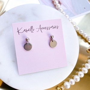 May include: A pair of silver stud earrings with a small round disc and a ball on top. The earrings are displayed on a pink card with the text "Kamelli Accessories".