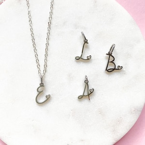 May include: A silver-colored necklace with a cursive letter "E" pendant and three individual letter charms: "L", "B", and "A". Each letter has a small heart detail. The jewelry is displayed on a white marble surface against a pink background.
