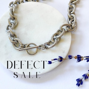Può includere: Una collana a catena argentata con chiusura a leva. La collana è su una superficie di marmo bianco con il testo "DEFECT SALE" sotto.