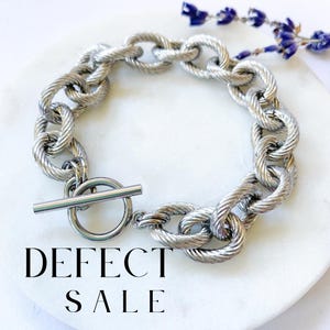 Puede incluir: Pulsera de cadena plateada con cierre de palanca. La pulsera está hecha de eslabones gruesos y retorcidos. El texto "DEFECT SALE" es visible en la imagen.
