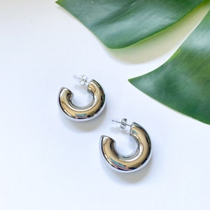 May include: A pair of silver hoop earrings with a thick, rounded design. The earrings are made of metal and have a polished finish.