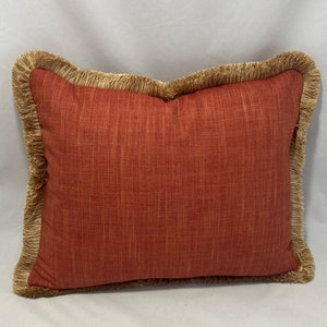 May include: Red throw pillow with a brown fringe border. The pillow is made of a textured fabric.