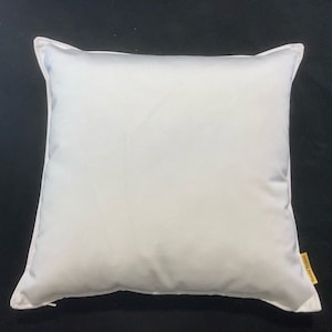 Puede incluir: Una almohada cuadrada blanca con una cremallera blanca en el borde inferior. La almohada está sobre un fondo negro.