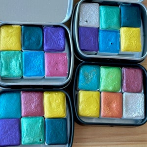 Four watercolor tins. Each tin includes six half pans with different colors of shimmering and glittering paint.