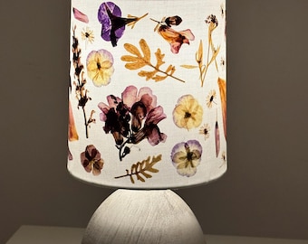 Peut inclure: Un abat-jour blanc avec un motif floral composé de fleurs séchées dans des tons de violet, rose, jaune et marron. L'abat-jour est sur un pied de lampe blanc.