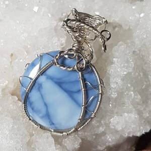 May include: A silver wire wrapped pendant featuring a blue and white opal gemstone. The pendant has a delicate, intricate design and is perfect for adding a touch of elegance to any outfit.