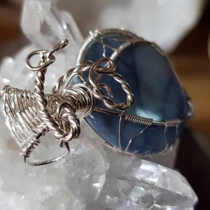 May include: A silver wire wrapped pendant featuring a blue agate stone. The pendant is displayed on a white crystal surface.