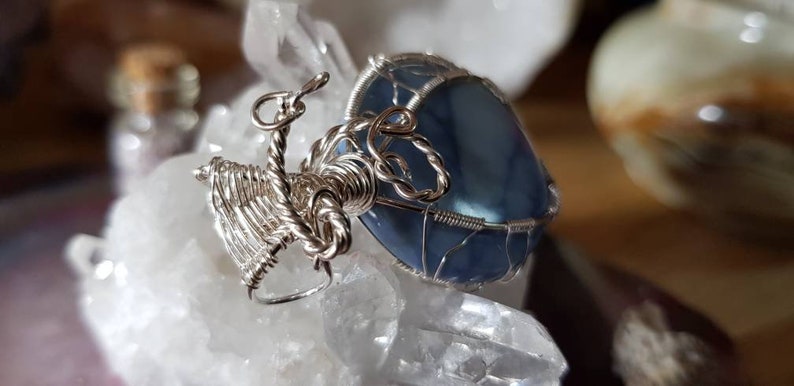 May include: A silver wire wrapped pendant featuring a blue agate stone. The pendant is displayed on a white crystal surface.