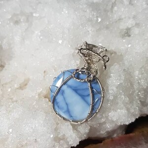 May include: A blue and white opal gemstone pendant wrapped in silver wire. The pendant is sitting on a white crystal surface.