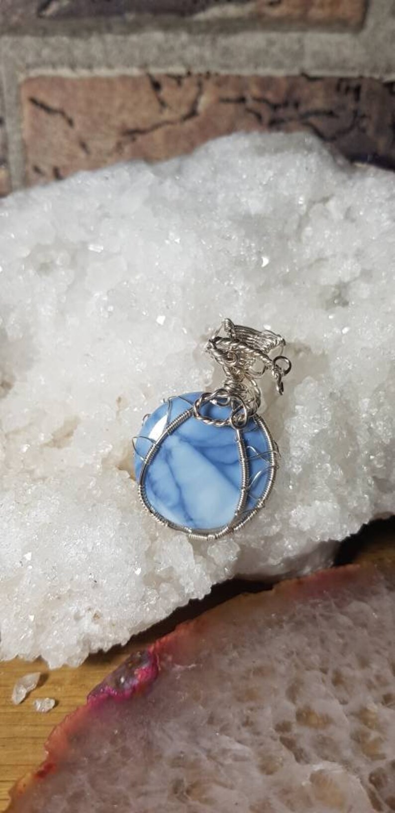 May include: A blue and white opal gemstone pendant wrapped in silver wire. The pendant is sitting on a white crystal surface.