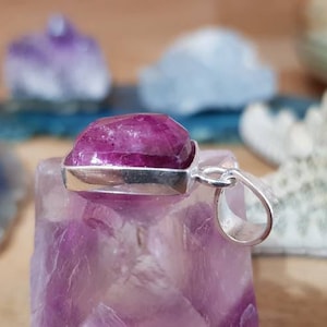 Puede incluir: Un colgante de piedra preciosa rosa con un engaste de plata. El colgante está sobre un cristal morado.