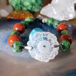 Puede incluir: Un brazalete hecho con piedras rojas, verdes y blancas. La pieza central es una gran flor de cuarzo transparente.