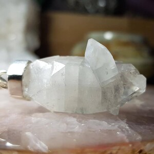 Może przedstawiać: Wisiorek z przezroczystego kryształu kwarcu z srebrną zawieszką. Kryształ jest fasetowany i ma unikalny, geometryczny kształt.