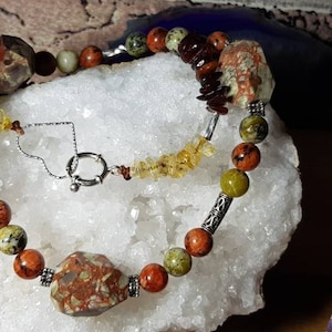 Puede incluir: Un collar de piedras preciosas multicolores con un cierre de plata. El collar presenta piedras verdes, naranjas y marrones.