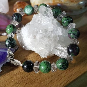 Puede incluir: Un brazalete hecho con cuentas verdes, negras y transparentes. El brazalete está sobre una superficie de madera con un cúmulo de cristales blancos en el fondo.
