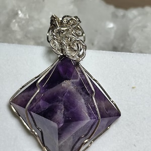 Puede incluir: Un colgante de cristal de amatista púrpura envuelto en alambre de plata. El cristal está facetado y tiene una forma geométrica.