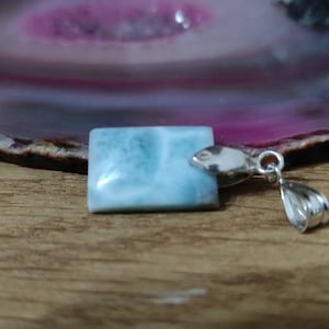 Puede incluir: Un colgante de piedra preciosa azul claro con una argolla de plata. La piedra preciosa tiene una apariencia nublada y veteada.